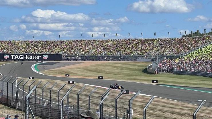 A wide view of the Landostand at Silverstone Circuit. Max Verstappen is shown driving his Red Bull in the foreground.