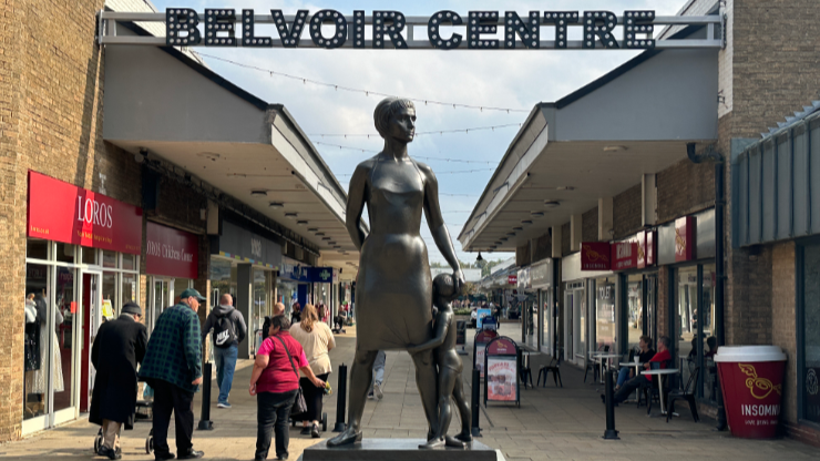 A large bronze statue of a woman in a dress, with a child hugging her leg in the foreground.  Shoppers can be seen in a precinct behind.