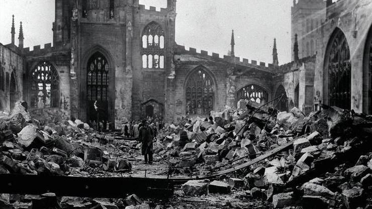 An old black and white photo showing inside a ruined cathedral with rubble strewn everywhere while the outer portion of the building remains standing. People can be seen entering the site and looking around at the scene.