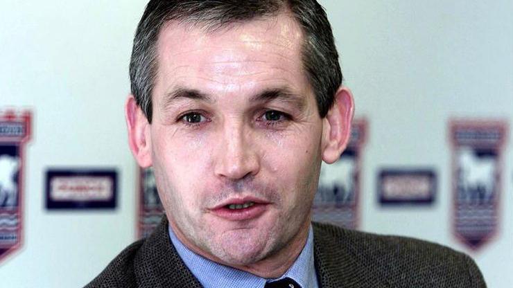 George Burley dressed in a tie, shirt and tweet jacket talking in front of a background of Ipswich Town badges