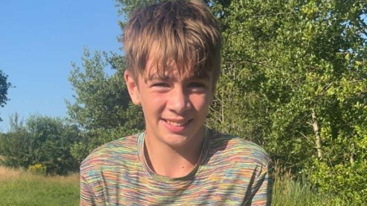 Logan smiles for the camera in front of a backdrop of trees. He has golden-coloured short hair and wears a stripey multi-coloured top.
