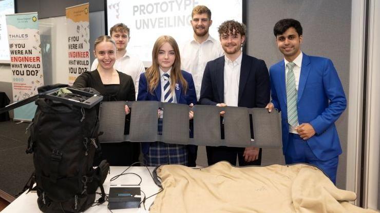 Rebecca in school uniform holding a prototype of electric backpack and blanket next to five men and one woman