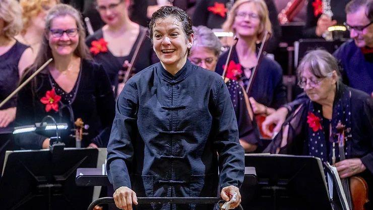 Natalia Luis-Bassa is standing in front of an orchestra. She has short dark hair and is wearing a mandarin-style black jacket. She is smiling and her hands are resting on a music stand. Behind her are several women with various string instruments in their hands and music stands in front of them. Each is wearing black clothes with a large red flower pinned to the right of their chests.