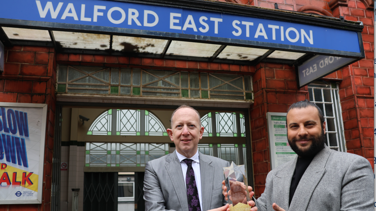 Jeremy Newmark is wearing a grey suit and stood near the entrance to Walford East Station as he passes an award to Chris Clenshaw