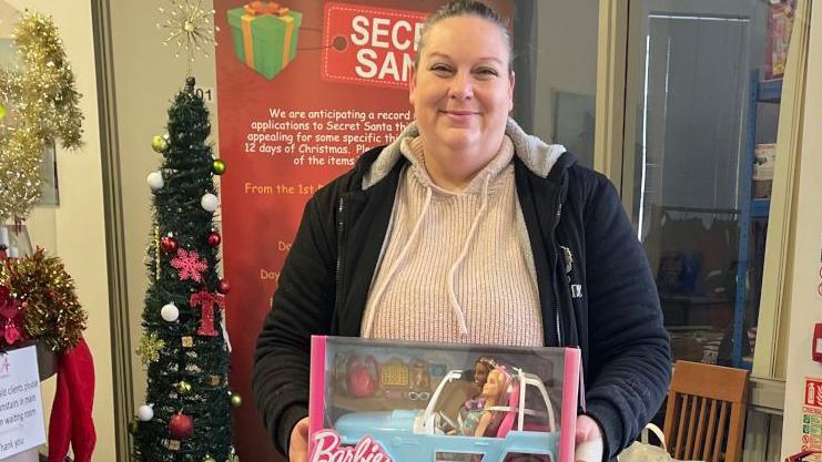 Ann Marie Allsopp holding a Barbie toy. She has a light coloured jumper on and dark jacket. Behind her is a Christmas tree