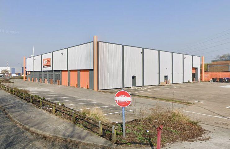 A pale grey warehouse photographed from the road. It looks empty, and there is graffiti on a road sign at the entrance to its car park. It has orange signage on it, and three orange garage doors along one of its sides. There is another garage door on another side of the building which looks to be open, and there are industrial bins in its car park. The grass borders are messy and unkempt.