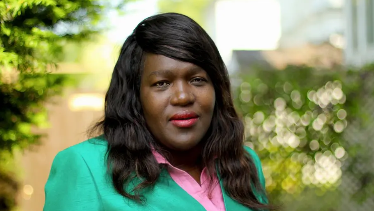 A woman with black hair looks at the camera. She is wearing a green blazer with a pink shirt beneath it. There are hedges behind her.