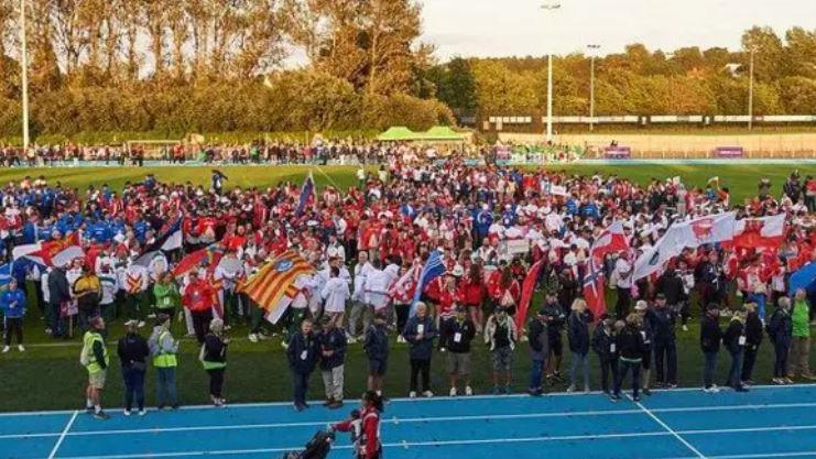 A crowd of people with flags lining a blue athletics track under the afternoon sun.