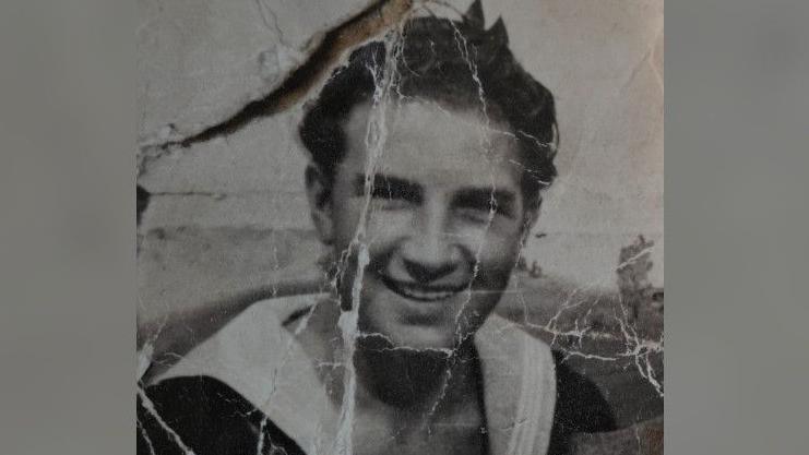 A black and white picture of a young man, He has short dark hair and is looking towards the camera, smiling. The photograph has white lines or creases on it.