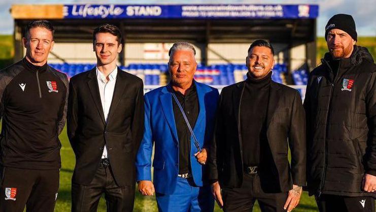 Barrie and Scott Drewitt-Barlow standing on the pitch at Maldon & Tiptree FC with other club members. A stand with blue seats is behind them. Barrie is wearing a blue suit and has slicked back hair. Scott is wearing a black suit with a black rollneck jumper and is smiling. The other people in the picture are looking at the camera while smiling, except one man who looks serious.