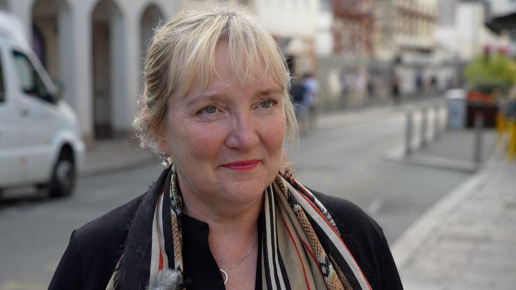 Tricia Warwick is wearing a scarf with a black blazer and has blonde hair tied back. She is stood on one side of a street with a road and a parked vehicle in the background. 