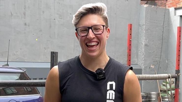 Laura Hollywood stands outdoors in front of a grey wall, wearing a sleeveless black shirt with white vertical text on the front.