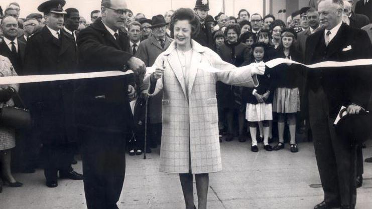 A black and white photo shows a woman in a large overcoat cutting a ribbon, with two gentlemen in smart suits and a bowler hat, with a crowd of onlookers of all ages behind them in smart dress and some in uniforms