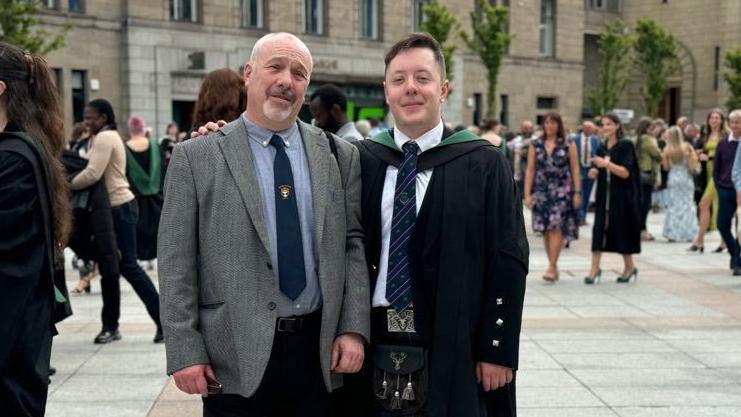 An older man wearing a grey blazer, navy tie and blue shirt stands next to a younger man in a graduation suit, also wearing a kilt