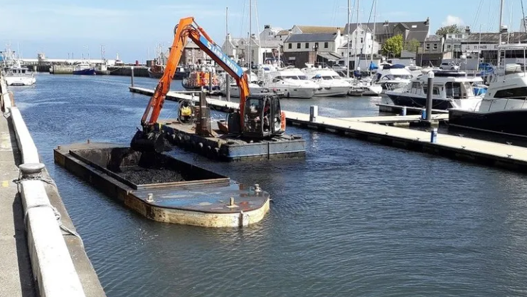 Silt is dredged from Peel marina with a digger on a barge lifting it onto a another barge for transportation. 