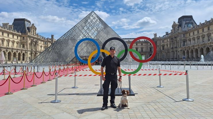 A police officer in all black clothes holds the lead of a brown and white dog, who is sat on the ground, in front of a statue of the five Olympic rings and the Louvre