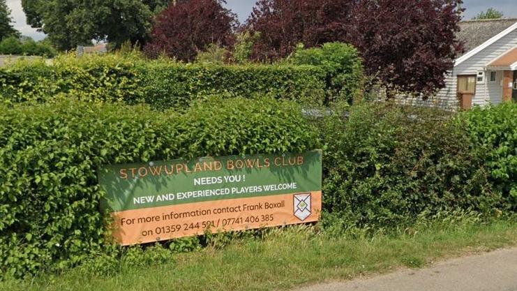 A sign for Stowupland Bowls Club in front of a hedgerow. 