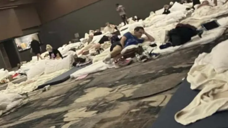 A slightly grainy image of a conference room - it has a brown patterned carpet and brown walls. There are rows of dark grey mattresses on the ground, with white sheets and pillows on top. People are sitting and lying on them, with a few also standing up.