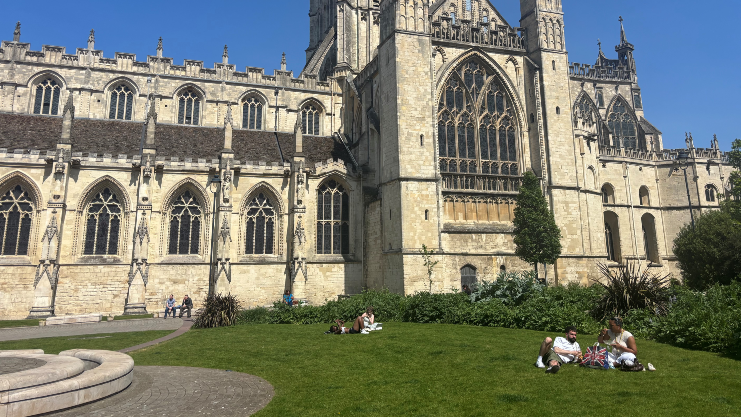 A photograph of a gothic cathedral taken on a sunny day. Two groups of people sunbathe in front.  