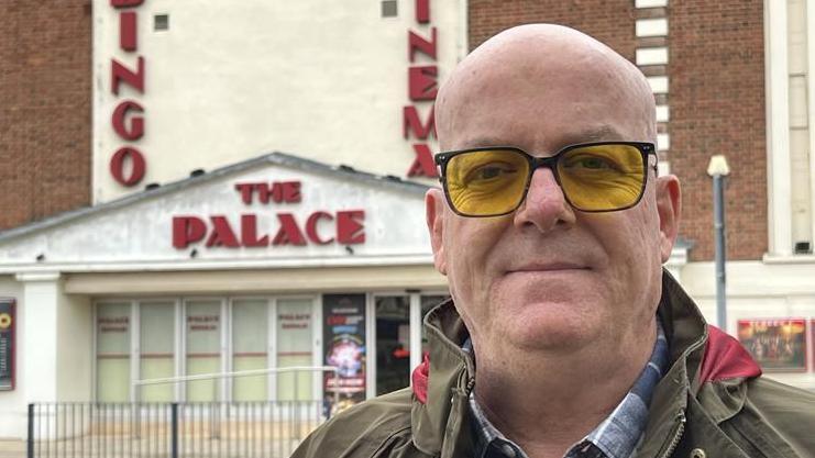 Mark Jones stands in front of the cinema. He is bald and wears glasses with yellow frames as well as a green coat and blue chequered shirt.