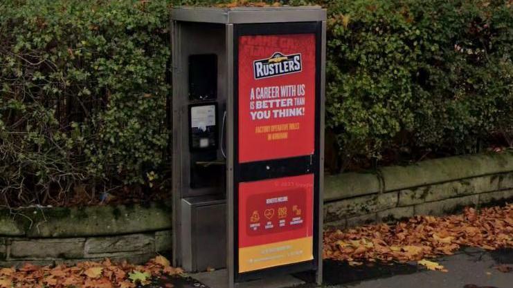 A phone box, which has one window obscured by a red poster containing an advert for Rustlers, with a hedge over a small wall in the background.