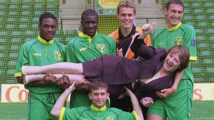 Delia Smith poses with Norwich City players at Carrow Road in July 1998 as the football club's new kit is revealed. Five players - four wearing green football kit and one in an orange and black shirt - hold Delia Smith horizontally. She is wearing a dark plum-coloured suit and white top and shoes.