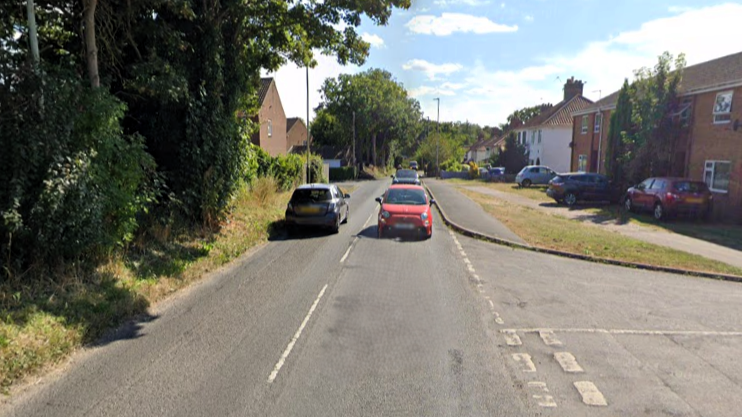 Cars driving along Hellesdon Road. Terrace houses line one side of the road and trees the other