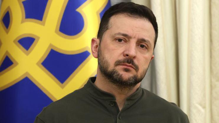 Volodymyr Zelensky looks over reporters during a press conference. The yellow and blue of the Ukrainian flag can be seen over his shoulder.