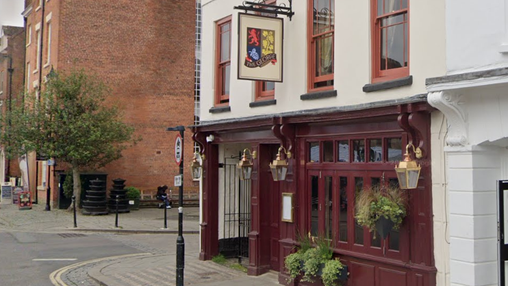A pub on a street corner with a ground floor that is maroon in colour. The top half of the building is white with orange window frames. There is a sign that reads "The George"