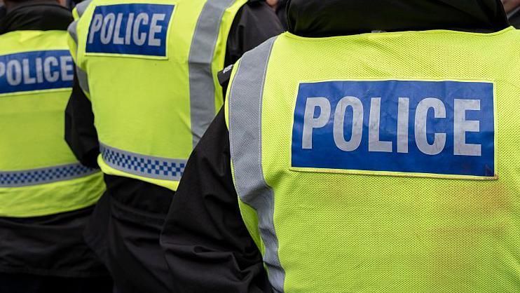 Stock photo of a police man facing away form the camera. He is wearing a black jacket with a yellow high vis jacket that reads POLICE on the back.