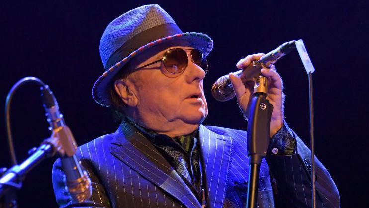 Image of Van Morrison wearing pinstripe navy suit singing into a microphone. He is wearing a bowler hat and tinted glasses.