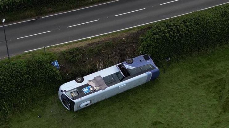A bus overturned on grass near a main road 