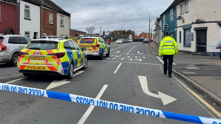 Two Essex Police cars are on the left of a road behind blue and white police tape. A section of the road has been cordoned off. A police officer is walking away from the camera on the right side of the road.
