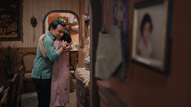 A still from the movie, which shows Norma's husband hugging Norma in the living room of their house. He is looking off to the side.
