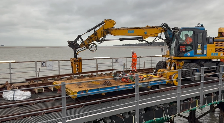 A yellow digger holding a steel girder on the railway tracks with two engineers wearing orange work overalls.