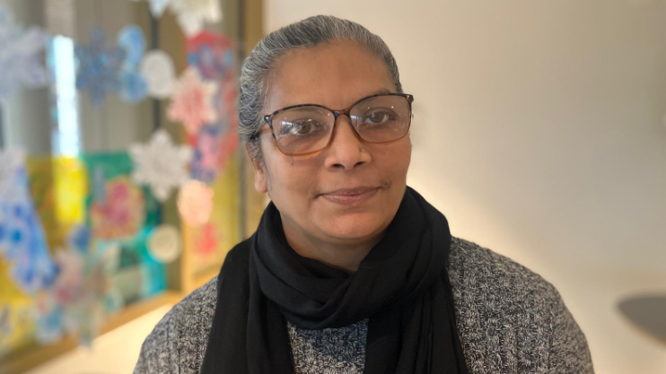 A woman with grey hair and glasses looks at the camera. She wears a black scarf and a grey jumper.