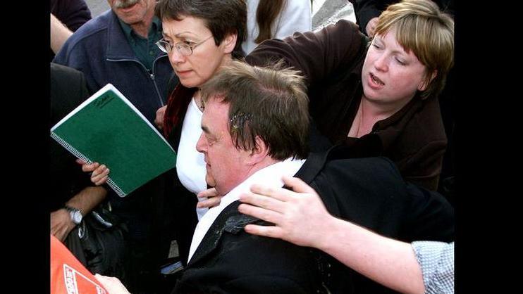 John Prescott covered in egg after being attacked during a visit to the North Wales seaside resort of Rhyl in May, 2001. He has egg yolk in his hair and a woman is reaching across the front of him with an arm in a protective manner. He is being ushered through a crowd of people.