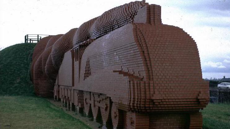 Brick Train pictured in 1999. It is made of brown bricks arranged in the form of a train. It is surrounded by low grass. 