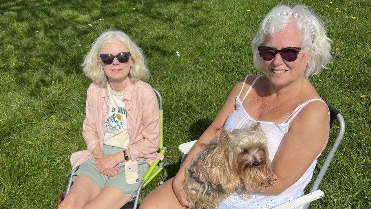 two women sat in garden chairs sunbathing. They both have white hair and wearing sunglasses. The woman on the right has a small dog on her lap.