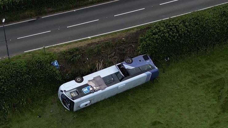 A purple and white bus is seen on is side in a field