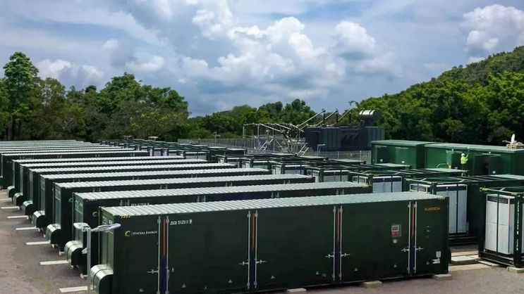 A row of about 20 green energy storage units about the size of a lorry container with trees around the site 