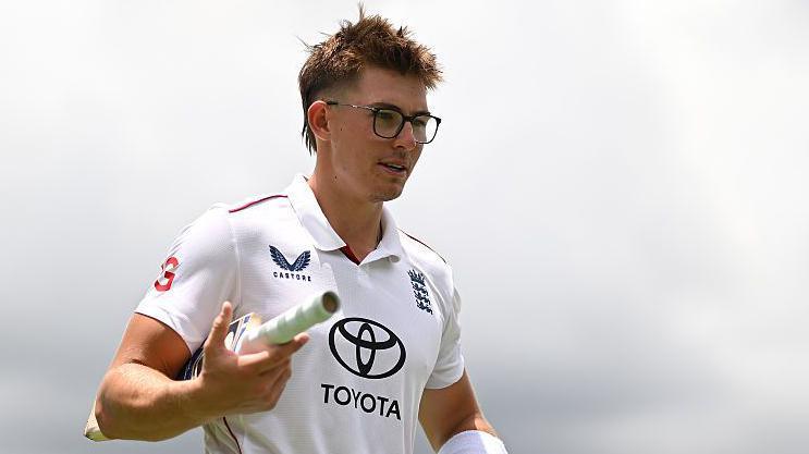 Asa Tribe of the England Lions walking off the field with a bat tucked in between his right arm.
