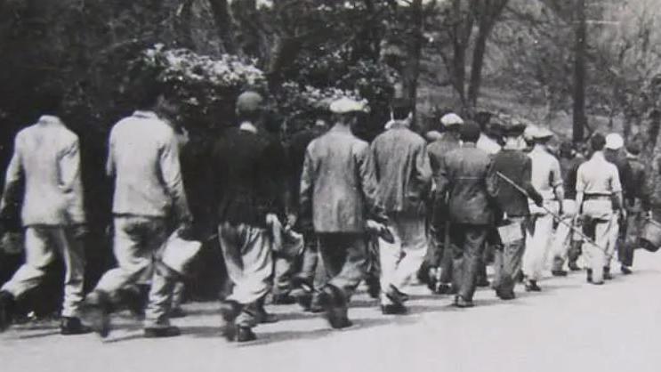 Black and white image of men dressed in dirty clothes and boots being marched along the road