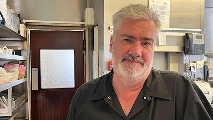 Ian Stead, who has a grey beard and wearing a black shirt, stands in the kitchen of his fish and chip shop