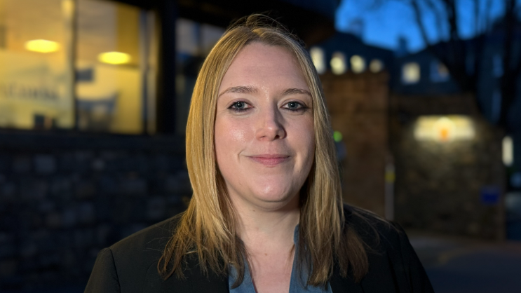 Poppy Murray in front a wall and a building. It is dark and the background is blurred.