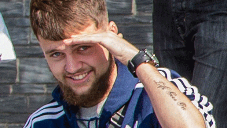 Conner, a young man with a beard and writing tattooed on his left arm, shields his face from the sun with one hand and smiles at the camera. He is wearing a blue jacket with three white stripes on the arms