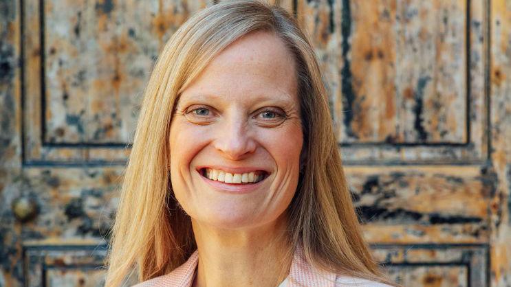 A woman with long blonde hair smiles widely as she stands in front of an old brown door.