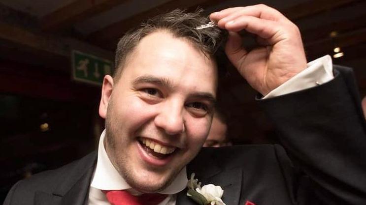 Michael Riddiough-Allen laughs while touching his hair at a wedding. He has a suit, red tie and flower in his lapel.
