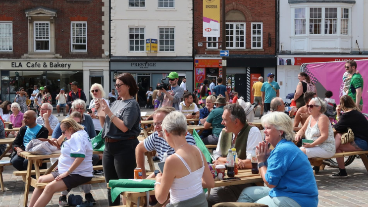 A large group of people in Northampton's Market Square at a fan zone. Some are standing up, others are sitting on benches, and there are shops behind them. 