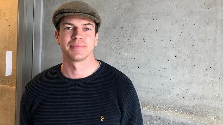 Ross Bonner stood in front of a wall wearing a flat cap and and dark jumper